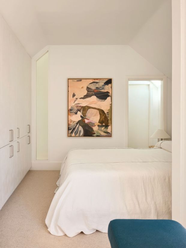 A bright, compact bedroom with a large white bed, integrated white built-in wardrobes, pale carpet, and a softly angled ceiling. An abstract artwork provides a muted pink, brown, and dark accent above the bed, while a teal upholstered chair sits in the foreground. A mirrored or reflective opening behind the bed adds depth to the otherwise minimal, calm interior.