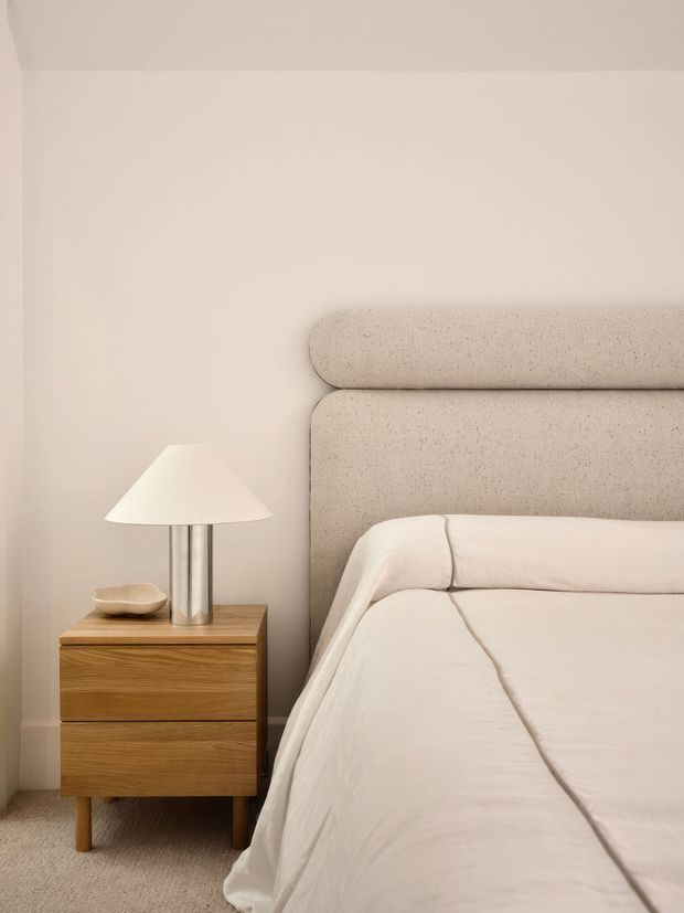 Close-up interior view of a calm, softly styled bedroom. A large upholstered, rounded-edge headboard in warm beige sits against a plain beige wall, beside a mid-tone wooden bedside table with drawers and tapered legs. A minimalist table lamp with a white fabric shade and metallic cylindrical base rests on the nightstand, while cream bedding fills the foreground. The palette is warm and neutral with subtle textured surfaces and gentle lighting.