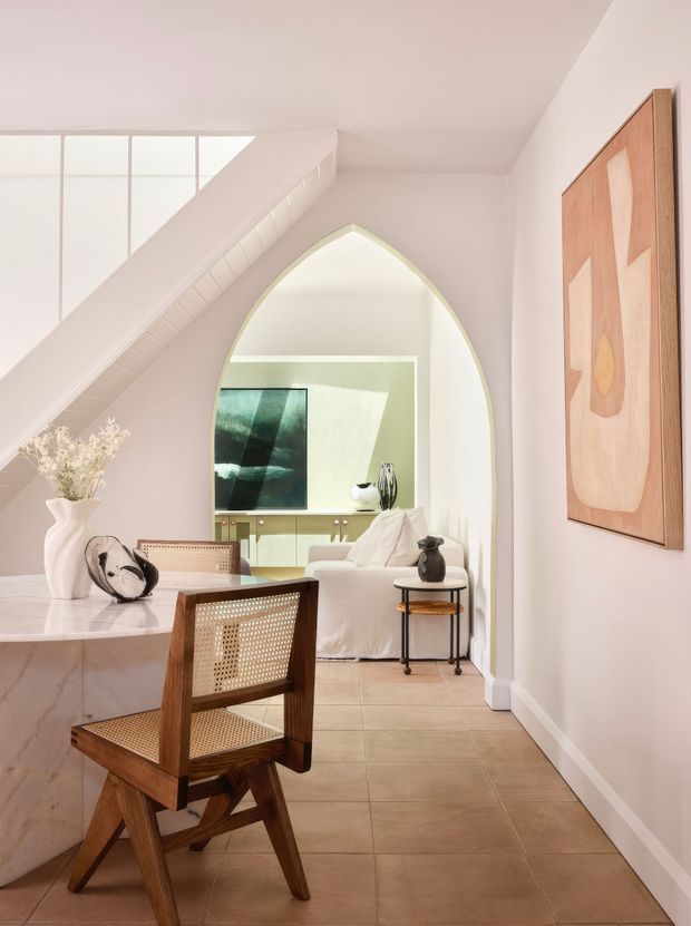 A refined open-plan interior viewed from a dining area beneath a sloping stair or ceiling structure. The foreground features a marble-topped dining table and mid-century wooden cane dining chairs. Beyond a distinctive pointed archway is a softly furnished living room with a white sofa, side table, built-in cabinetry and large artwork. The palette is light and warm, with tiled flooring, pale walls, and natural timber accents.
