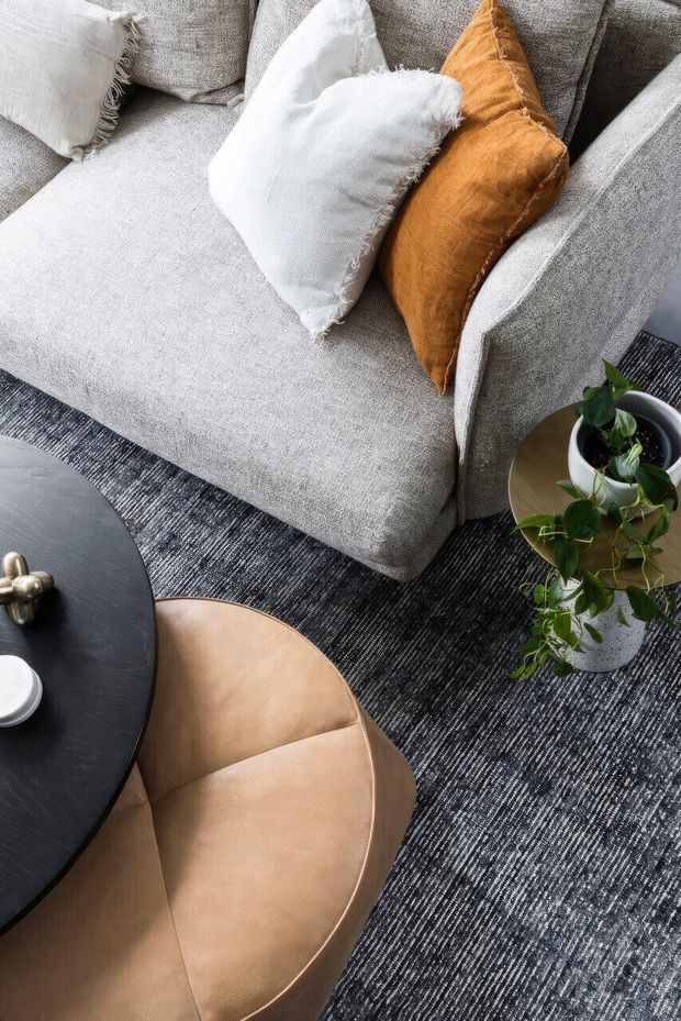A close-up overhead view of a contemporary living room seating arrangement, featuring a light grey upholstered sofa with white and ochre cushions, a tan leather lounge chair, a round black coffee table, a textured dark patterned rug, and a potted plant.