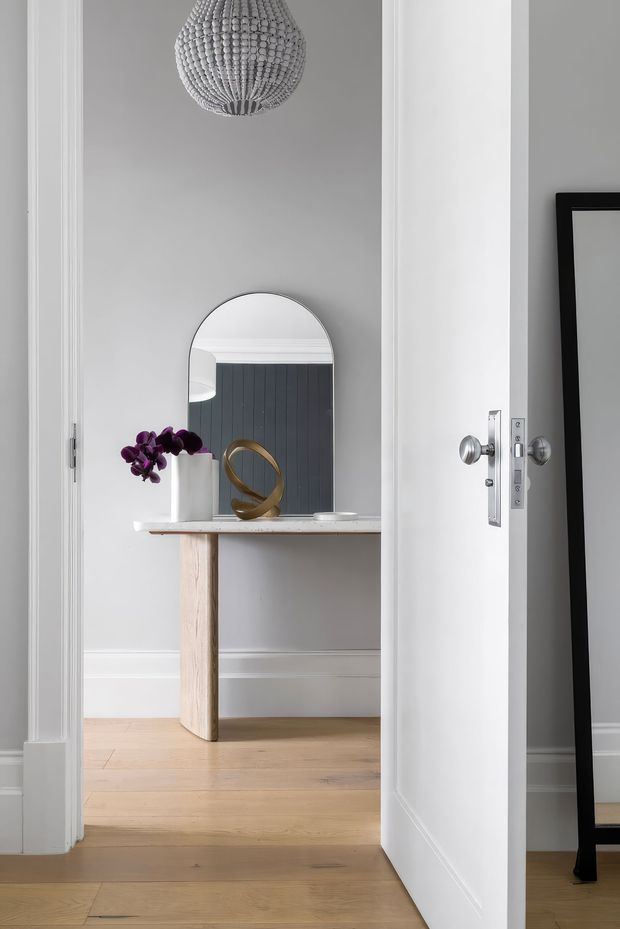 A refined interior view through an open white-trimmed doorway into a small hall or entry area. The space features pale timber flooring, soft grey walls with white moulded skirting and architraves, a slim console table, an arched mirror, sculptural decor, a purple orchid, and a textured pendant light. A large black-framed mirror is partially visible in the foreground.