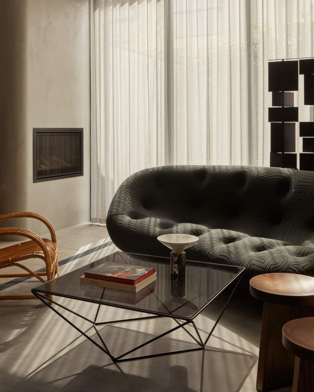 A sophisticated living room flooded with warm natural light through full-height sheer curtains. The space features a large dark sculptural tufted sofa, a geometric black metal and glass coffee table with books, rounded wooden side tables, woven wooden lounge chairs, a wall-mounted fireplace, and a striking black-and-white geometric artwork or screen. The palette is neutral with strong contrast and a refined, textural, mid-century-inspired feel.