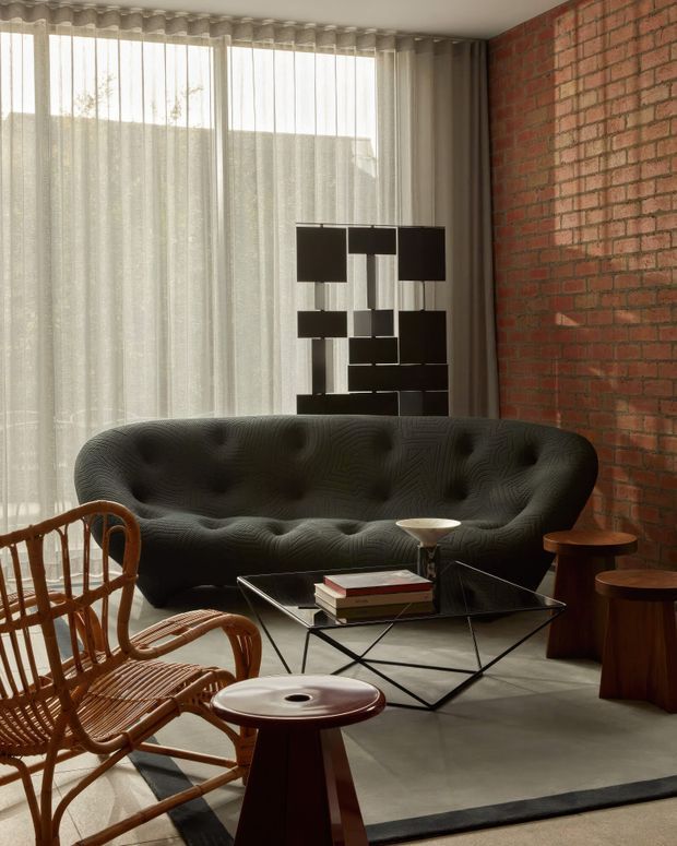 A refined mid-century-inspired living room with a deep charcoal tufted sofa, geometric black coffee table, sculptural pendant light, woven rattan lounge chairs, and timber side tables. Full-height sheer curtains diffuse daylight along one side, while an exposed red brick wall adds industrial character. The room has polished concrete flooring and a restrained, warm-neutral palette.