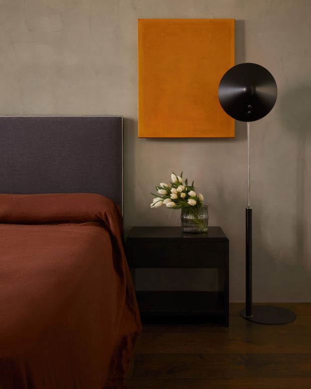 A moody, refined bedroom vignette with a dark upholstered bedhead, rust-brown bedding, dark timber flooring, and a black bedside table. A tall black floor lamp stands beside the bed, while a warm orange artwork and a vase of white flowers provide contrast against the grey-beige wall.