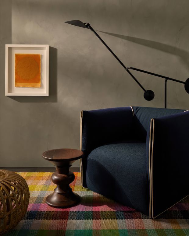 A moody, richly styled living room vignette with a deep navy upholstered armchair, sculptural dark timber side table, long black articulated floor lamp, framed orange abstract artwork, and a multicoloured checkered rug. The room has a textured grey plaster wall and strong directional lighting that casts dramatic shadows. A woven basket or chair is partially visible in the foreground.