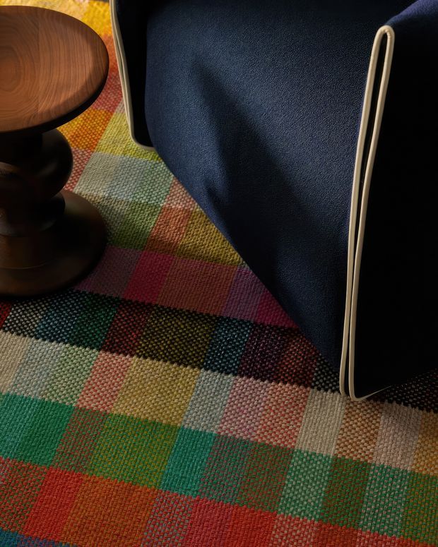 Close-up interior detail showing a richly multicoloured, geometric woven rug beneath a dark navy upholstered chair with a slim light-coloured metal frame, alongside a dark stained timber pedestal side table. The composition focuses on tactile textile patterns, saturated colours and furniture details.