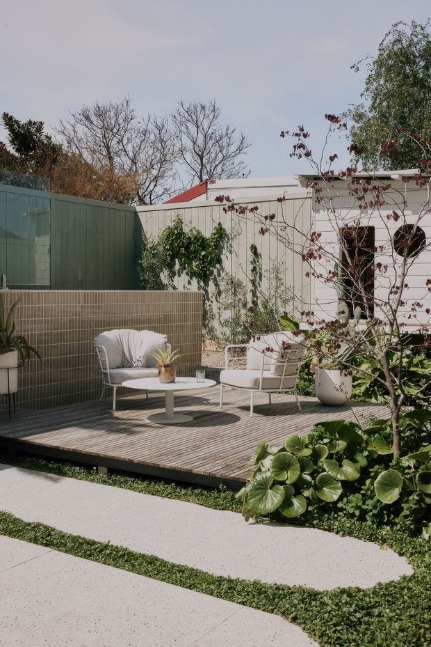 A landscaped outdoor courtyard or patio with a raised timber deck, a tiled privacy wall, pale outdoor lounge chairs, and a small round table. The space is bordered by planted garden beds, a concrete stepping path, fences, and a light-colored pavilion or building structure. The setting has a calm, contemporary garden-lounge character with muted natural tones.
