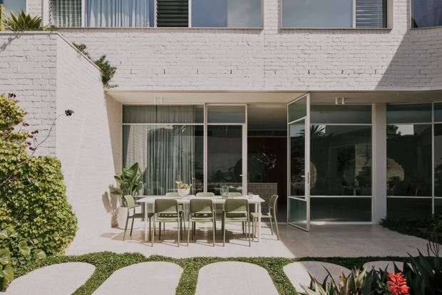 A contemporary two-storey residential exterior with a white-painted brick facade, large aluminium-framed glazed doors and windows, and a ground-level outdoor dining terrace. The terrace opens onto a landscaped garden with curved stepping stones, clipped planting, succulents, and a boundary wall. Strong sunlight creates crisp shadows across the facade and patio.