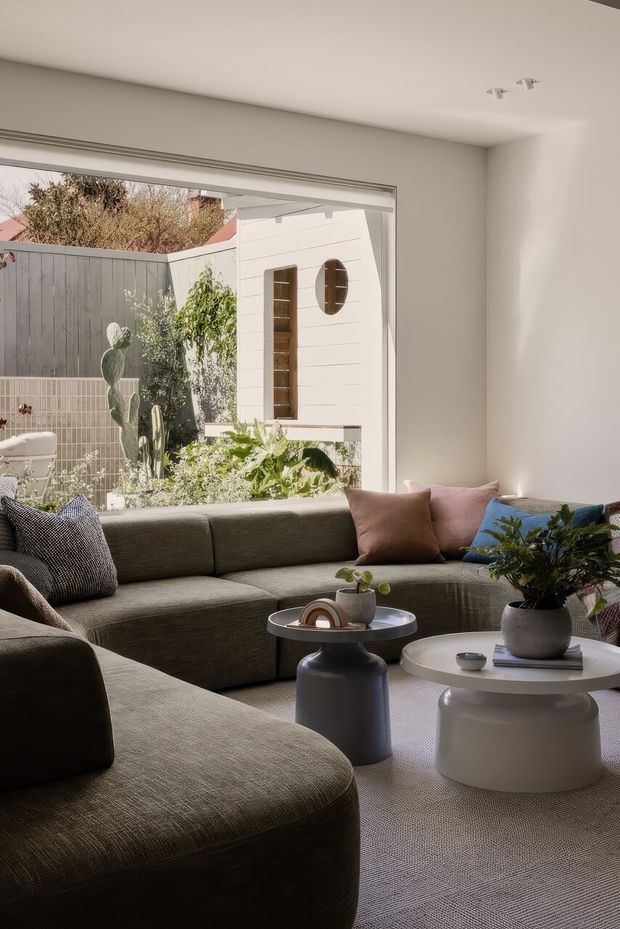 A softly lit contemporary living room with a large olive-grey modular sectional sofa, pale neutral walls, and a broad glazed opening overlooking a planted courtyard. Two rounded coffee tables, sculptural ceramic vessels, cushions, and indoor plants create a calm, refined atmosphere. The courtyard includes a white-clad garden structure and mature planting.