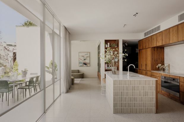 A bright contemporary open-plan kitchen and living space with a large tiled island, integrated timber cabinetry, pale stone benchtop, and full-height glazed doors opening to a leafy outdoor terrace. The room features light terrazzo-style flooring, white walls, a neutral palette, and a built-in dining area visible through the glazing.