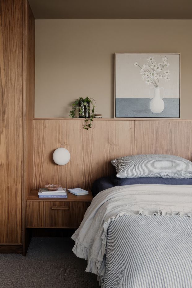 A warm, refined bedroom detail featuring a full-height timber wall and integrated timber headboard cabinetry, a built-in bedside table with a round wall light, layered neutral bedding, and framed floral artwork. The room has a calm, earthy palette and soft contemporary styling.