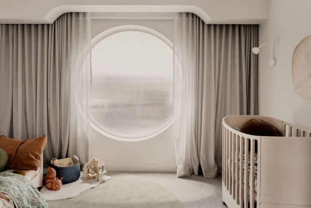 A softly lit nursery with warm neutral tones, floor-to-ceiling taupe curtains, sheer drapery, and a distinctive large circular window. A curved white crib occupies the right side, while a cushioned seating area with pillows, toys, and a small playhouse sits on a pale rug to the left. The room has rounded architectural detailing and a calm, refined atmosphere.