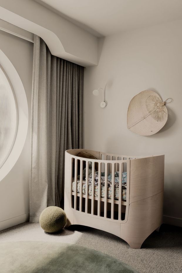 A softly lit nursery with a curved light-wood baby cot, neutral textured carpet, muted green rug, full-height taupe curtains, and warm off-white walls. The room features a rounded architectural opening, a small wall sconce, and woven circular wall art, creating a calm, refined contemporary setting.