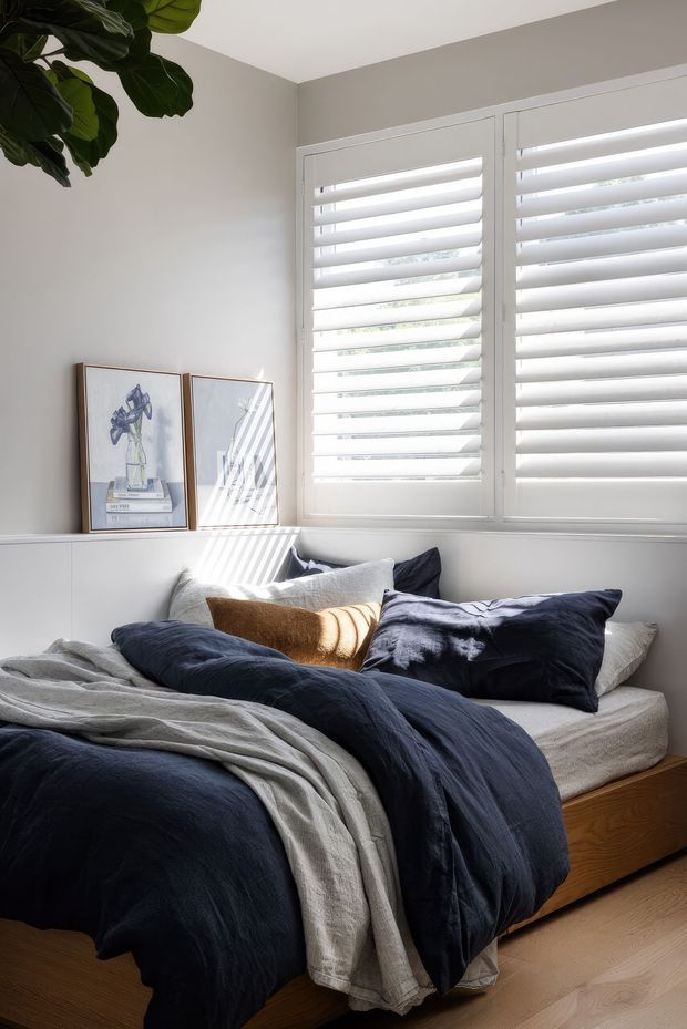 Bright, calming bedroom with a low timber bed, layered navy and light grey linen bedding, neutral walls, white plantation shutters, and framed botanical artwork. Sunlight filters through the shutters, creating striped shadows across the bed and wall. A large leafy indoor plant adds a natural touch.
