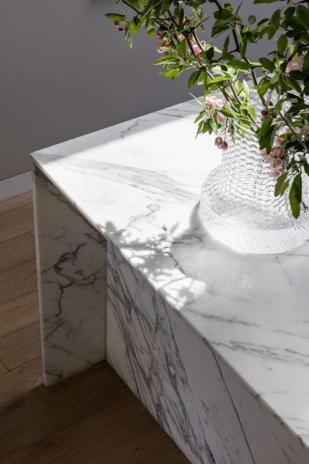 Close-up architectural detail of a substantial white marble island or counter with prominent grey veining, waterfall-style side panels, and a softly rounded edge. The counter sits against a neutral grey wall above warm timber flooring. A textured clear glass vase with leafy flowering branches adds a natural decorative element, while strong sunlight casts foliage shadows across the marble.