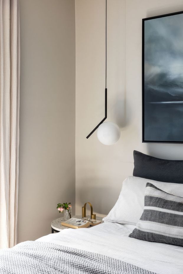 A softly styled bedroom corner with a neatly made bed, layered white and charcoal bedding, a round bedside table with small decor, a sculptural black wall light, floor-length curtains, and framed artwork. The neutral palette and tactile textiles create a calm, refined atmosphere.