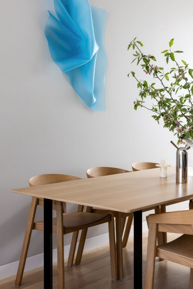 A softly lit dining area with a light timber dining table and matching wooden chairs, set against a pale grey wall and timber floor. A large sculptural translucent blue wall artwork provides a bold focal point, while leafy flowering branches in a tall metallic vase add a natural element to the tabletop.