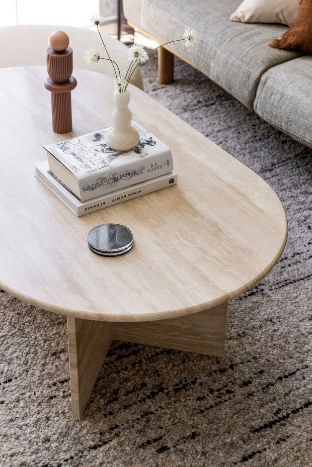Close-up interior view of a softly styled living room, centred on a large rounded oval coffee table with a pale beige stone or travertine-like finish and matching angular pedestal legs. Books, a small vase of white flowers, and a ribbed brown decorative object sit on the tabletop. A grey upholstered sofa, textured neutral rug, cushions, and warm natural materials create a calm, contemporary setting.