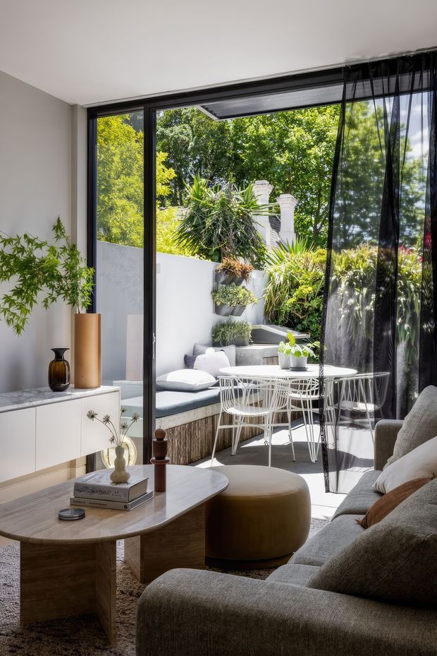 Bright contemporary living room opening onto a leafy, landscaped courtyard through large black-framed glazed doors. The interior features a substantial neutral sectional sofa, rounded timber coffee table, upholstered ottoman, white sideboard, indoor plants, and soft black sheer curtains. Outside is a compact tiered courtyard with built-in bench seating, white outdoor dining furniture, raised planters, and lush planting.