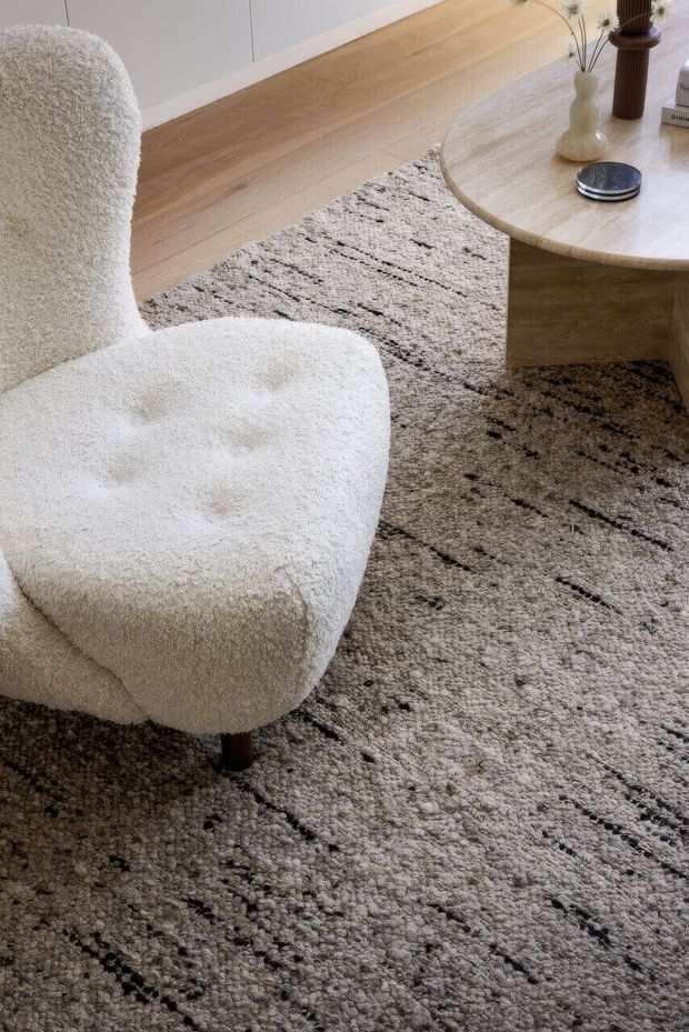 Close-up interior view of a softly styled living room, featuring a sculptural cream boucle armchair, a textured light grey rug with irregular dark linear detailing, and a rounded light timber coffee table. Neutral cabinetry or wall panelling is visible in the background, with small decorative vessels and a smart device on the table. The overall look is warm, calm, tactile, and contemporary.