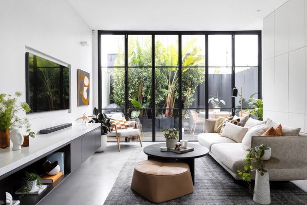 Bright contemporary living room with a large cream sectional sofa, round black coffee table, leather ottoman, wall-mounted television and sleek low media joinery. Full-height black-framed glazing opens visually to a leafy courtyard, while white wall cabinetry and polished concrete-like flooring create a clean, refined interior.