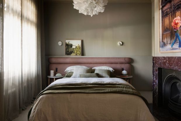 A refined bedroom with a low upholstered bed and rounded mauve headboard, layered neutral bedding, and matching bedside tables. Soft daylight filters through full-height curtains on the left, while a dark green-grey wall, sculptural pendant light, framed artwork, and an ornate marble fireplace create a warm, elegant atmosphere.