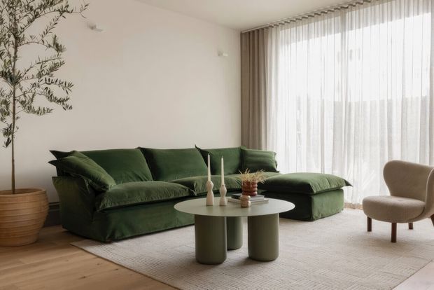 A softly lit contemporary living room with a deep forest-green sectional sofa, round sculptural coffee tables, a neutral textured area rug, light timber flooring, sheer full-height curtains, and a sculptural beige armchair. A potted indoor tree adds an organic element.