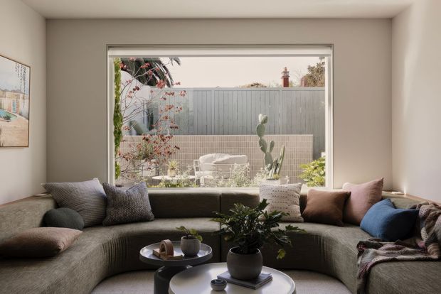 A warm, contemporary living room centred on a large corner-like picture window overlooking a private courtyard. A deep curved sectional sofa fills the foreground with layered cushions, a throw, round coffee tables, and potted plants. The neutral interior contrasts with the planted outdoor space and grey fence beyond.