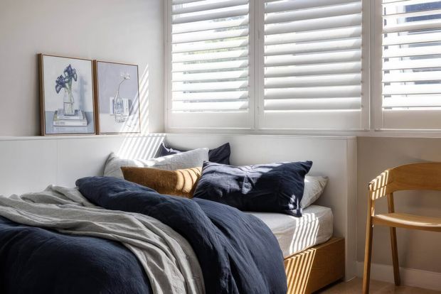 Bright, compact bedroom with a bed layered in navy and grey bedding, white built-in cabinetry, framed botanical artwork, plantation shutters, and a simple timber chair beside the window. Strong sunlight creates striped shadows across the bed and floor, giving the room a calm, inviting feel.