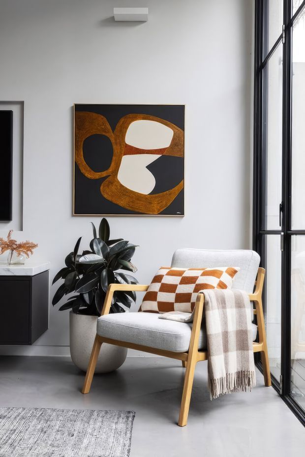 A bright, refined sitting corner with a light upholstered timber armchair beside full-height black-framed glazing. The space features a grey concrete-look floor, white walls, an abstract artwork in black, cream and ochre, a dark-leaf potted plant, and warm patterned textiles. A partial dark built-in cabinet and rug are visible at the edges.