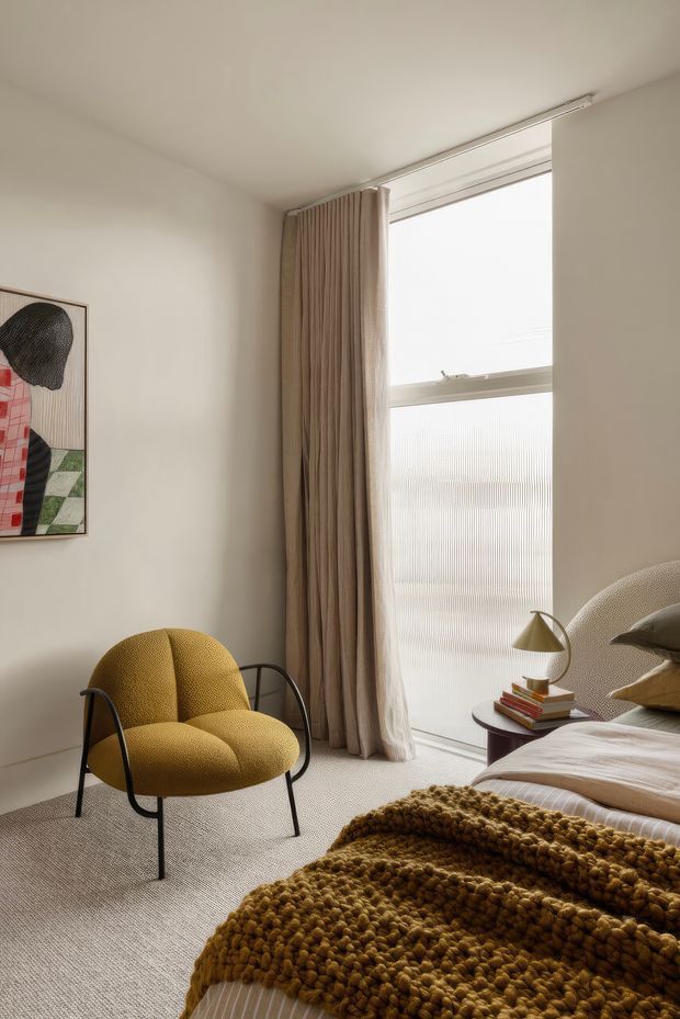A softly lit, contemporary bedroom with warm neutral walls, a tall frosted-glass window dressed in full-height beige curtains, and textured carpet flooring. The room features a mustard-yellow upholstered lounge chair with black metal arms, a partially visible upholstered bed with layered neutral bedding, a small dark side table with books and a brass-toned table lamp, and a chunky mustard knitted throw. Abstract framed artwork adds a graphic accent.