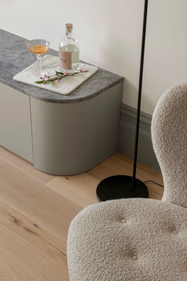 A softly styled interior corner featuring a curved grey built-in cabinet or sideboard with a dark stone-look benchtop, pale timber flooring, and a textured cream boucle armchair. A tray with a bottle, cocktail glass, and small flowers sits on the cabinet, beside a slim black floor lamp. The overall composition is warm, minimal, and refined.