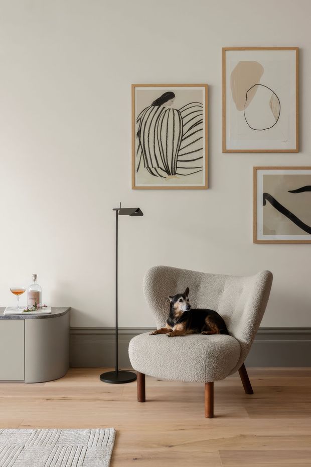 A softly styled living room with a sculptural cream boucle armchair, a small black dog, a slender black floor lamp, a curved side cabinet with a drink, framed abstract and botanical artwork, warm timber flooring, and grey-beige wall panelling. The space has a calm, refined, contemporary feel with natural light and muted neutral tones.