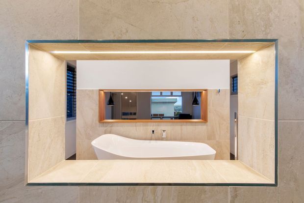 A refined contemporary bathroom viewed through a large rectangular wall opening framed in dark metal. The space features full-height beige stone-look wall finishes, a freestanding white bathtub, a wide illuminated mirror with timber-toned trim, recessed lighting, and a calm neutral palette. A reflected view reveals additional bathroom fixtures and glazing.