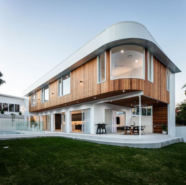 Exterior photograph of a contemporary two-storey residence with a distinctive curved roofline and rounded upper-level corner. The facade combines vertical timber cladding, white rendered surfaces, extensive glazing, and a covered outdoor entertaining area. A lawn, patio, and pool edge are visible in the foreground.