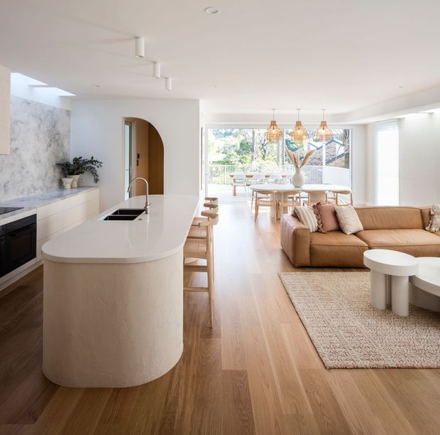 Bright open-plan contemporary interior combining a kitchen, dining area and living room. The kitchen features a large rounded island with a white stone benchtop, pale cabinetry, integrated appliances and a marble-look feature wall. Warm timber flooring extends through the space. The living area includes a tan sectional sofa, neutral textured rug and round white coffee tables, while the dining area is lit by three pendant lights and opens to a bright outdoor terrace through extensive glazing.