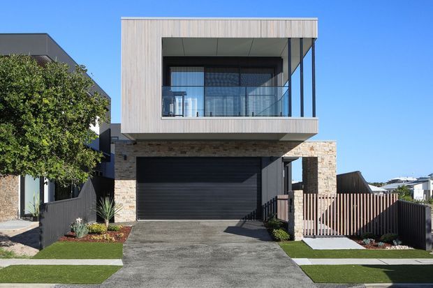 Street-facing exterior of a contemporary multi-storey home with a projecting upper-level box, enclosed balcony, vertical light-coloured cladding, dark framing and garage door, and a contrasting textured masonry base. The front driveway, garage, fencing and landscaped verge are visible under clear daylight.