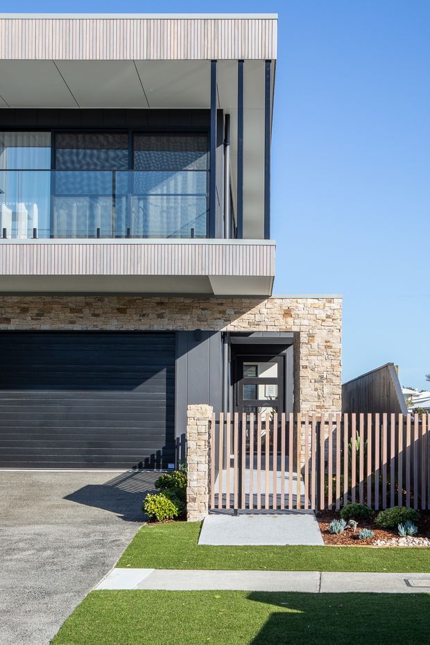 Street-facing view of a contemporary multi-storey residential facade. The house features pale vertical cladding, expansive dark-framed glazing, a projecting upper balcony with vertical balustrades, a natural stacked-stone garage surround and entry wall, a dark sectional garage door, and a timber-slatted pedestrian gate. The frontage includes a paved driveway, concrete path, and neatly landscaped lawn.