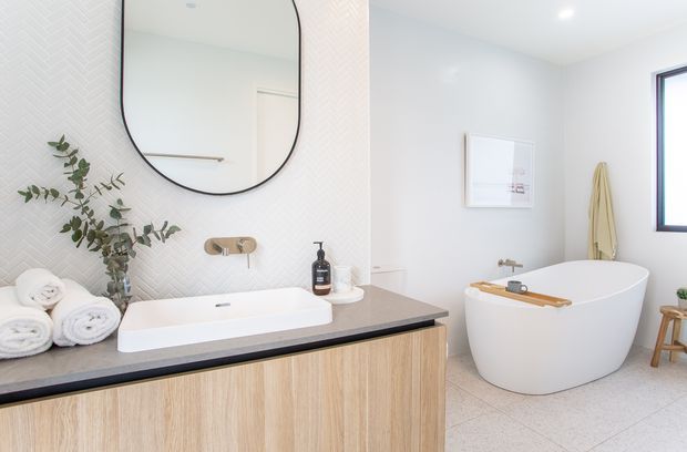 Bright contemporary bathroom featuring a floating timber-look vanity with a dark stone benchtop, integrated white basin, brushed brass wall-mounted tapware, and a freestanding white bathtub. The walls include subtle light herringbone-patterned tiling, with a large rounded black-framed mirror, pale floor tiles, recessed ceiling lighting, a black-framed window, and minimal accessories.
