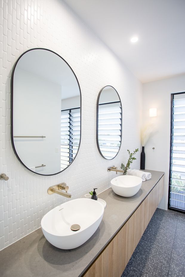 Bright contemporary bathroom featuring a long double vanity with two white vessel basins, a grey stone-look benchtop, warm timber-grain cabinetry, large arched mirrors, brass tapware, and white vertically patterned wall tiles. Louvered windows and a dark speckled floor add contrast.