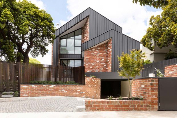 Street-facing exterior view of a contemporary multi-level residence with dark vertical metal cladding, exposed red brick feature walls, large glazed openings, timber fencing, and an integrated lower-level garage or carport opening. The home sits within a landscaped urban setting and uses strong geometric roof and facade forms.