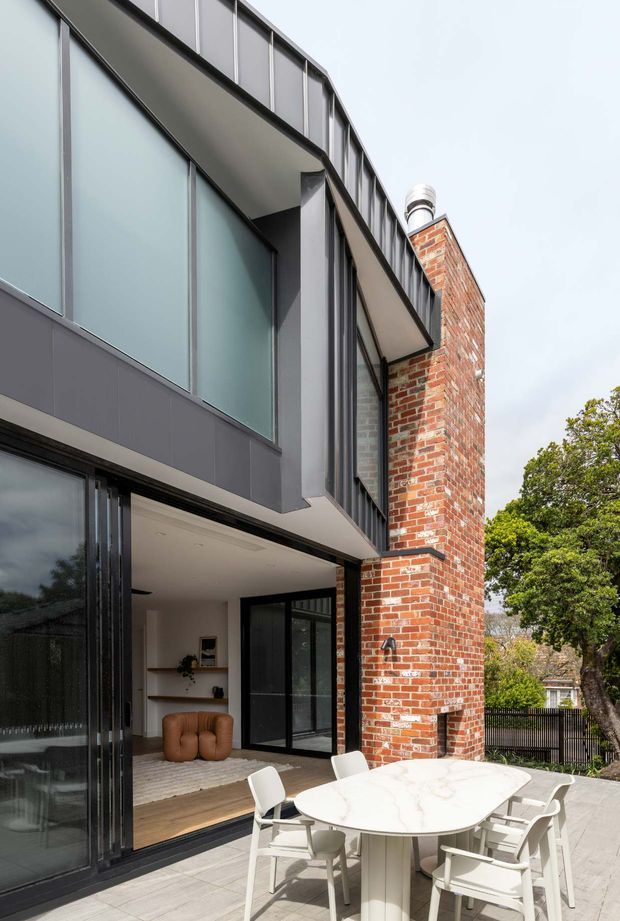 Contemporary two-storey residential extension viewed from an outdoor terrace. The facade combines large frosted or translucent-looking glazed panels, dark metal-framed glazing and cladding with an exposed red brick wall and chimney. An open-plan interior is visible through the folding glass doors, while the foreground contains a pale stone outdoor dining table and chairs on a paved terrace.
