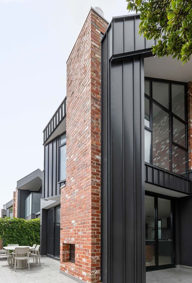 Exterior architectural photograph of a contemporary multi-storey residential building, combining an exposed red brick party wall with dark vertical metal cladding, large black-framed glazing, projecting balconies and a paved outdoor dining area. The facade appears to integrate an older brick structure with a modern addition.