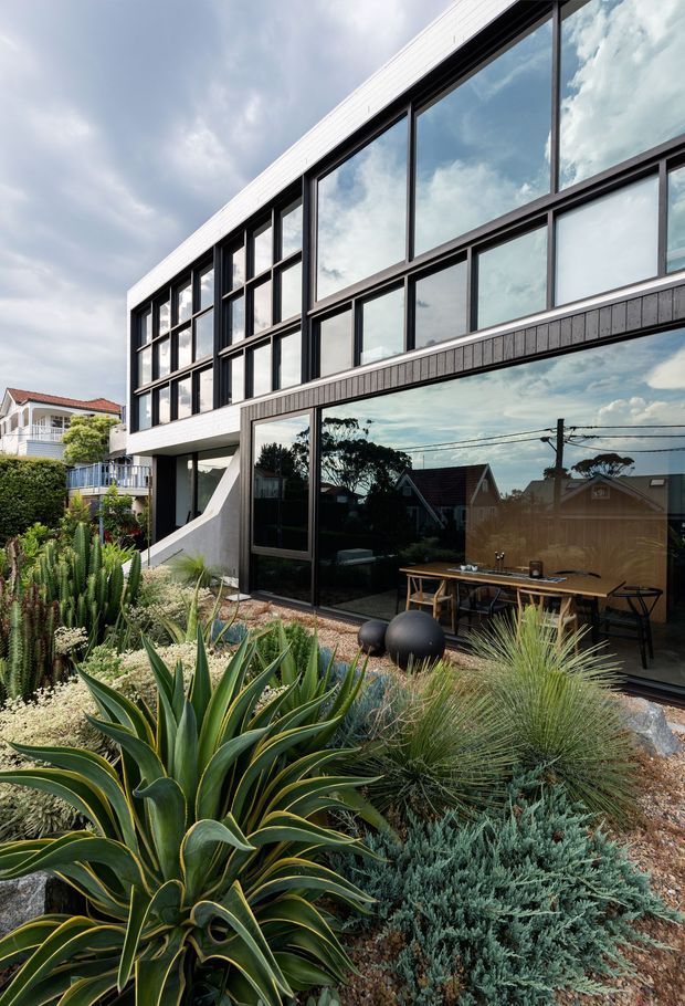 Exterior view of a contemporary multi-storey residence with a strongly rectilinear form, extensive floor-to-ceiling glazing, dark window frames, pale upper cladding and a textured dark lower facade. The building opens onto a landscaped outdoor dining terrace, with drought-tolerant architectural planting including agave, cacti and grasses. Reflections in the glazing show neighbouring suburban homes and overhead services.