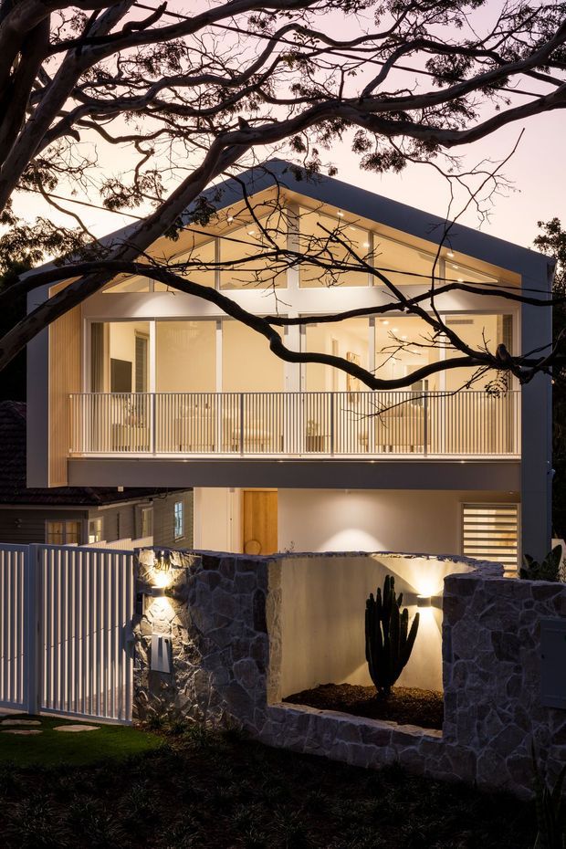 Dusk exterior view of a contemporary multi-storey residence set among mature trees. The home features a strong gabled roof form, extensive upper-level glazing, a broad balcony with slender vertical metal balustrades, warm interior lighting, and a lower-level entry screened by pale stone walls and a metal gate. Dark landscaping and silhouetted branches frame the illuminated facade.