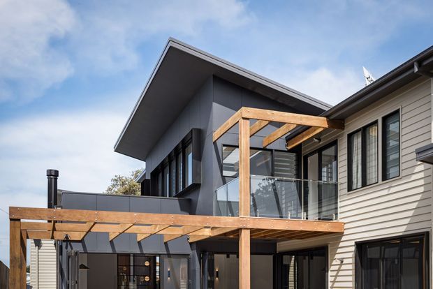 Exterior view of a contemporary multi-level residential extension featuring dark vertical cladding, light horizontal weatherboard siding, extensive glazing, and a timber-framed balcony or terrace with clear glass balustrades. The building has a prominent asymmetrical pitched roof and a covered timber pergola structure.