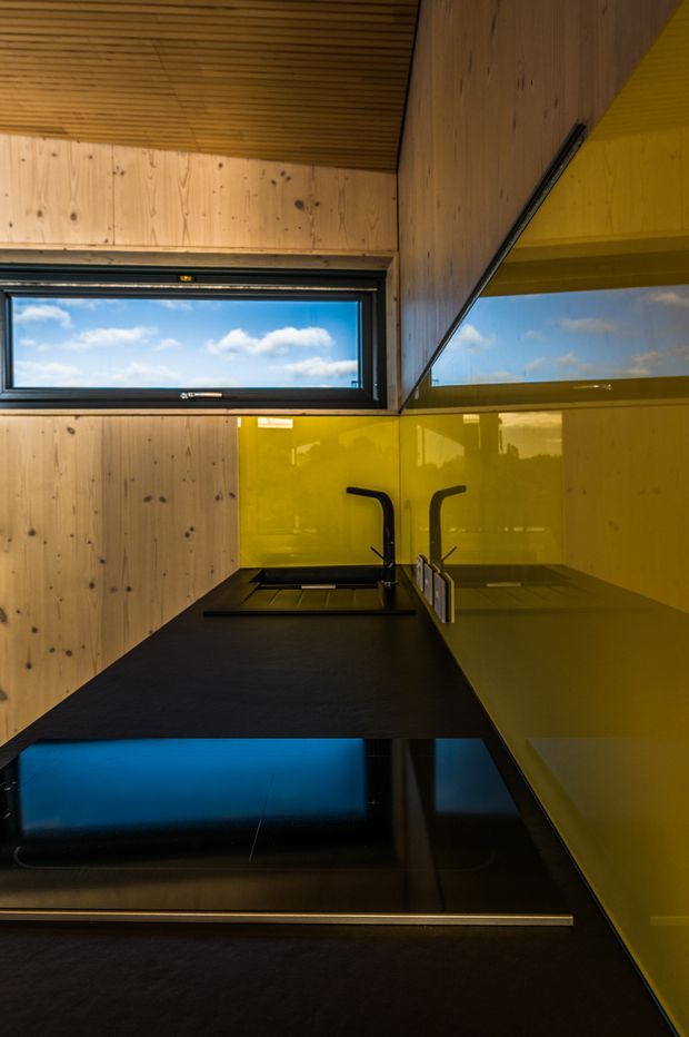 Compact modern kitchen with warm exposed timber wall and ceiling linings, a glossy vivid yellow backsplash and cabinetry, and dark black benchtops. A horizontal awning window brings in daylight and reflects the blue sky. The kitchen includes a black sink and angular black tap, with a strong high-contrast, contemporary colour scheme.