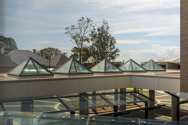 Exterior architectural view of a contemporary flat-roof building with a long glazed roof structure featuring repeated triangular pyramid skylights. A glass-covered lower-level walkway or atrium is visible in the foreground, with neighbouring pitched-roof buildings and mature trees beyond.