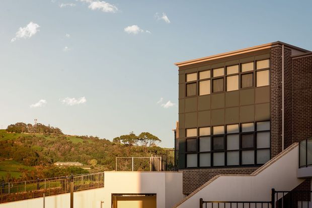 Exterior architectural view of a contemporary multi-storey building on a sloped site. The facade combines dark brickwork with large gridded window panels and dark metal framing, while terraces, stairways, and railings occupy the foreground. A green, hilly landscape is visible beyond the building.