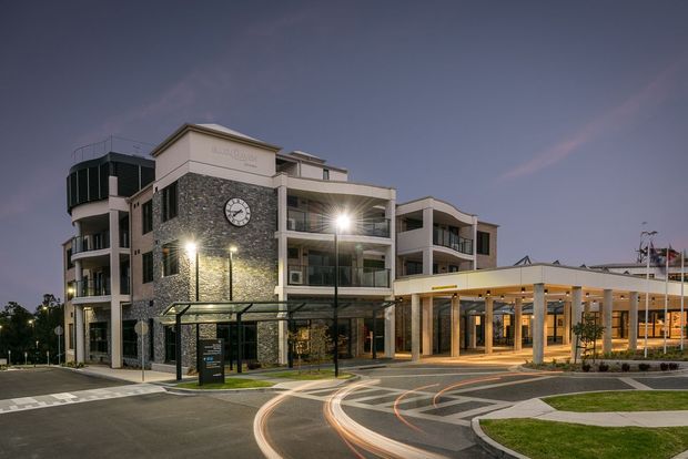 Dusk exterior view of a contemporary multi-storey commercial or civic building with a prominent dark stone-clad clock tower, balconies, extensive glazing, and a broad covered entrance canopy. Curved driveway markings and illuminated landscaping frame the forecourt.